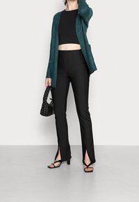 Cardigan lavorato a maglia verde acqua, top corto nero, pantaloni neri a spacco e sandali neri con cinturini. Borsa a tracolla nera intrecciata come accessorio.