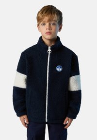 Niño vestido con una chaqueta de forro polar azul marino con rayas blancas en las mangas y un logo circular de North Sails en el pecho, de pie frente a un fondo liso.