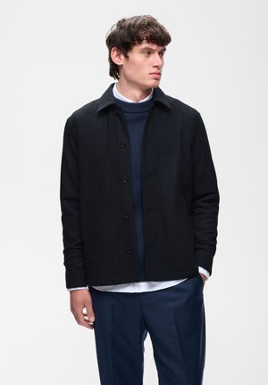 Ung mand iført en sort knappet jakke over en navyblå sweater og hvid skjorte, stående med den ene hånd i lommen og kigger til siden.