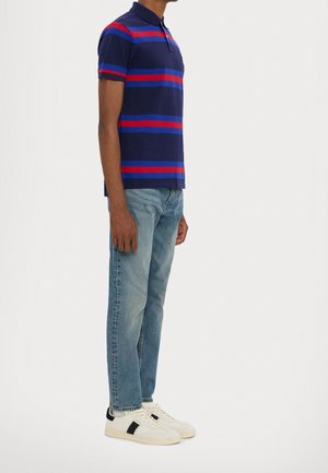Mann trägt blaues Polohemd mit roten und blauen Streifen, hellblaue Jeans und weiße Sneaker mit schwarzen Akzenten, steht seitlich vor weißem Hintergrund.