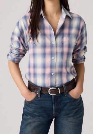Femme portant une chemise à carreaux pastel avec boutons et manches retroussées, un jean bleu et une ceinture marron foncé texturée avec une boucle argentée.