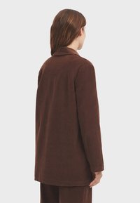 Blazer de pana marrón con cuello con muesca, corte recto y mangas largas. Presenta una tela texturizada con estrías visibles.