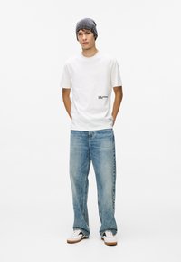 Balts T-krekls ar apkakli, uz kura priekšpuses ir loja, zili brīvi pieguloši džinsi, pelēks adīts beanie, un baltas sporta apavi ar melniem akcentiem un gumijas zolēm.