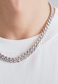 Collar de cadena de plata hecho de eslabones entrelazados, con un acabado pulido. Lleva puesto sobre una camiseta blanca de cuello redondo con cuello acanalado.