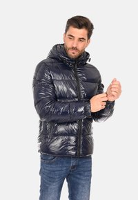 Giacca puffer blu navy con texture lucida, con zip frontale, cappuccio regolabile e polsini elasticizzati, indossata sopra dei jeans blu.