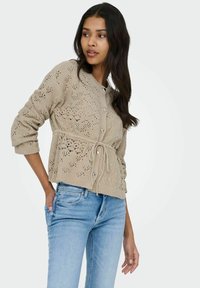 Beežist kootud kardigan keeruka pitsmustriga, hõbedaste nuppude ja vöökohale seotud detailiga, kombineeritud helesiniste teksadega.