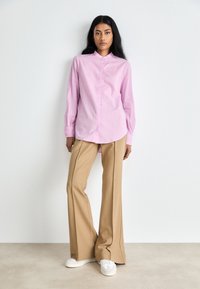 Camicia rosa con colletto alla mandarina, abbinata a pantaloni beige a gamba larga e sneakers bianche. Tessuto morbido e vestibilità sartoriale.