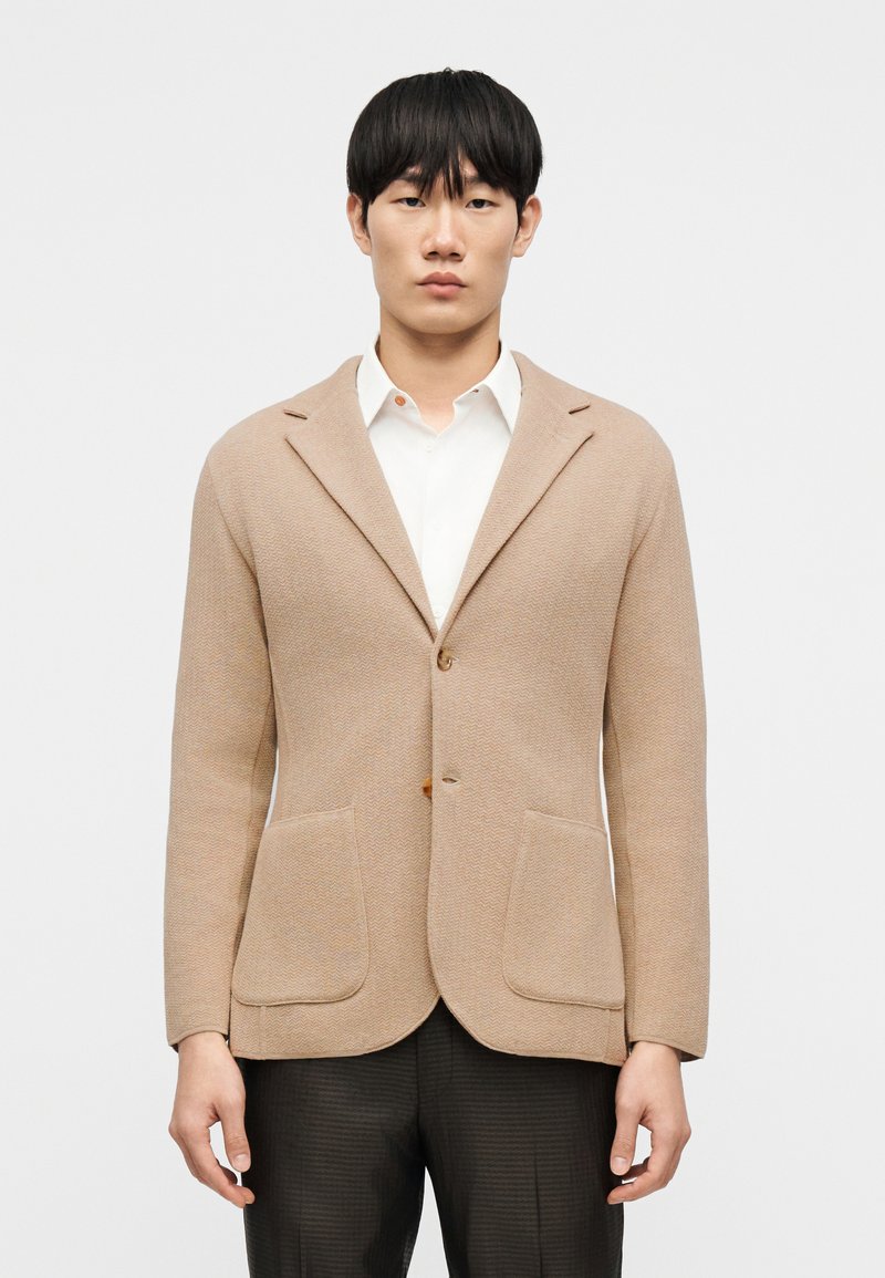 Μπεζ πλεκτό σακάκι με ελαφρώς υφασ μένο φινίρισμα, σχεδιασμένο με γιακά με χαραγή, δύο μπροστινές τσέπες και κλείσιμο με ένα κουμπί.