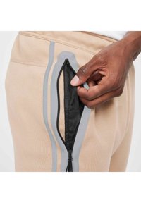 Pantaloni sportivi beige con strisce grigie presentano una tasca laterale con zip. La tasca ha un interno nero ed è aperta con una cerniera liscia.
