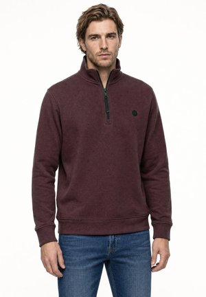 Mann trägt einen bordeauxfarbenen Pullover mit Viertel-Reißverschluss und blaue Jeans, steht vor einem weißen Hintergrund.