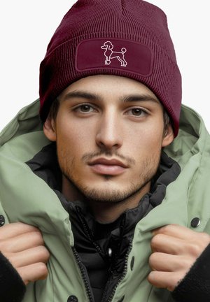 Bordeauxfarbene Strickmütze mit strukturierter Oberfläche und einem Stoffpatch, das eine weiße Umrisszeichnung eines Hundes zeigt. Trägt man zu einer grünen Parka-Jacke.