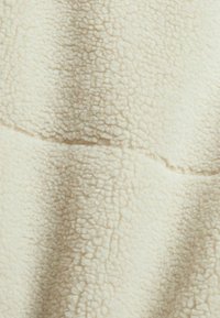 Kankaan fleece väri off-white, tekstuuripinta pehmeän ja pörröisen ulkonäön kanssa. Saumayksityiskohdat ovat näkyvissä, luoden osioita materiaalissa.