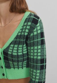 Zielony kardigan w kratę z czarnymi i jaśniejszymi zielonymi wzorami, dekoltem w serek i czarnymi guzikami. Wykonany z miękkiego, teksturowanego materiału.