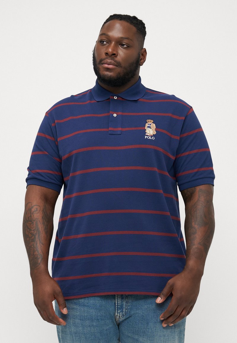 Marineblauwe polo met rode horizontale strepen, geborduurd berenlogo op de borst, kraag en korte mouwen. Stof van een katoenmix.