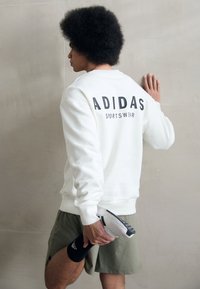 Balta džemperis ar melnu uzrakstu "ADIDAS SPORTSWEAR" aizmugurē, apvienots ar olīvu krāsas šortiem un turot baltu sporta apavu.