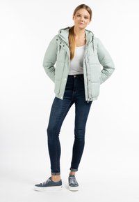 Giacca imbottita verde chiaro con cappuccio, chiusure a zip e a bottone, e tasche laterali. Indossata sopra una maglietta bianca e abbinata a jeans skinny scuri e sneakers blu.