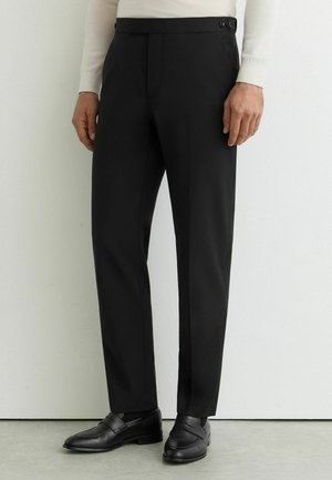 Mężczyzna w dopasowanych czarnych eleganckich spodniach z bocznym detalem zapinanym na guziki, w połączeniu z czarnymi mokasynami i białą koszulą z długim rękawem.