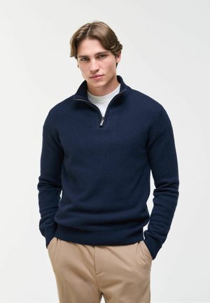 Giovane uomo con capelli castano chiaro che indossa un maglione blu navy con cerniera a quarto sopra una camicia bianca e pantaloni beige, in piedi con le mani nelle tasche.