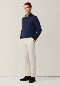 Sweater bleu marine à zip avec des poignets côtelés, associé à un pantalon beige clair et des chaussures foncées. Le tissu semble lisse avec un design classique.