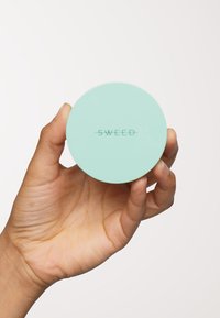 Un récipient rond de couleur menthe, avec une surface lisse, arborant le nom de la marque "SWEED" en relief dans une police minimaliste, tenu dans une main.