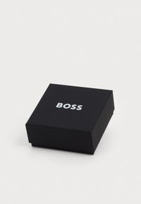 Cutie neagră din carton cu forma pătrată, având un logo alb "BOSS" pe suprafața superioară. Fără modele sau texturi suplimentare vizibile.