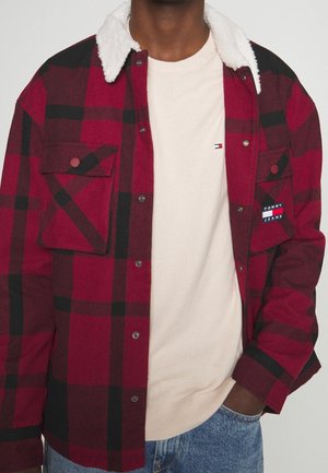 Mann trägt eine rot-schwarz karierte Jacke mit weißem Fleece-Kragen über einem beigen Pullover und blauen Jeans, Hand in der Tasche.