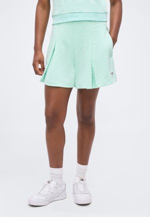 Egy személy alsó testtája, aki világoszöld pliszírozott rövidnadrágot, fehér térdzoknit és fehér Adidas tornacipőt visel egyszínű háttér előtt.