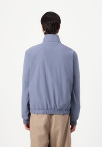 Homme portant une veste bleu clair avec manches et ourlet élastiqués, vu de dos, assortie à un pantalon beige sur un fond blanc uni.