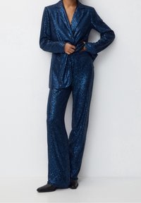Niebieska marynarka z cekinami i pasujące spodnie o dopasowanym kroju. Tkanina jest błyszcząca, z gładką teksturą i długimi rękawami.