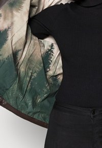 Wnętrze brązowej skórzanej kurtki zdobi wzór zielonego i beżowego lasu; na zewnątrz widoczny jest czarny prążkowany golf i ciemne spodnie.