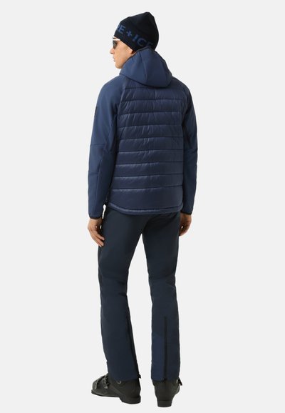 Marineblaue isolierte Jacke mit Kapuze, gesteppter Vorderseite und glattem Rücken, kombiniert mit marineblauen Hosen mit Reißverschlüssen an den Knöcheln und schwarzen Stiefeln.