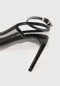 Sandale noire à talons hauts en cuir verni élégant, avec un panneau latéral en maille et une fine sangle de cheville maintenue par une boucle métallique.