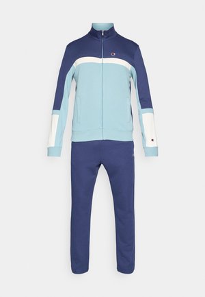 Tweedelig trainingspak met een blauwe en lichtblauwe ritssluitingjack, met witte en marineblauwe accenten, en bijpassende marineblauwe broek. Gemaakt van gladde stof.