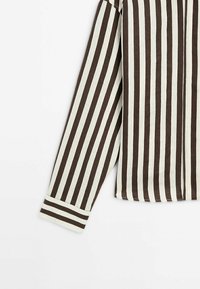 Chemise à manches longues avec des rayures verticales noires et blanches et un poignet à rayures horizontales sur un fond blanc.