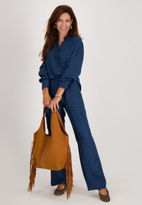 Marineblauwe jumpsuit met lange mouwen, knoopsluiting en elastische taille. Bruine tote bag met franjes en studs. Schoenen met luipaardprint.