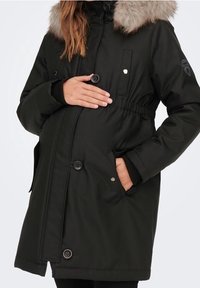 Svart isolerad parkas med huva av konstgjord päls, insvängd midja, knapprad och sidofickor. Mjuk textur och varm design.