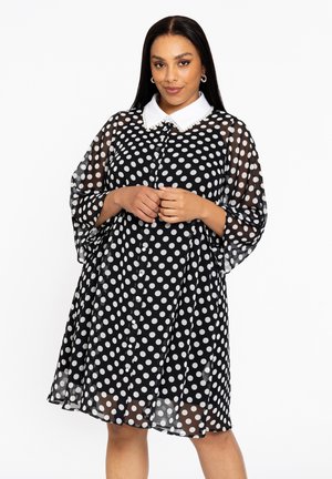 Robe noire à pois blancs avec un col blanc, des manches courtes et un design fluide. Fabriquée en tissu transparent avec des détails de boutons sur le devant.
