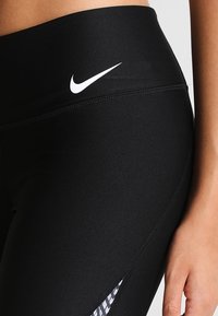 Czarne legginsy sportowe o dopasowanym kroju, z białym logo Nike na pasku w talii i wzorzystym panelem bocznym.