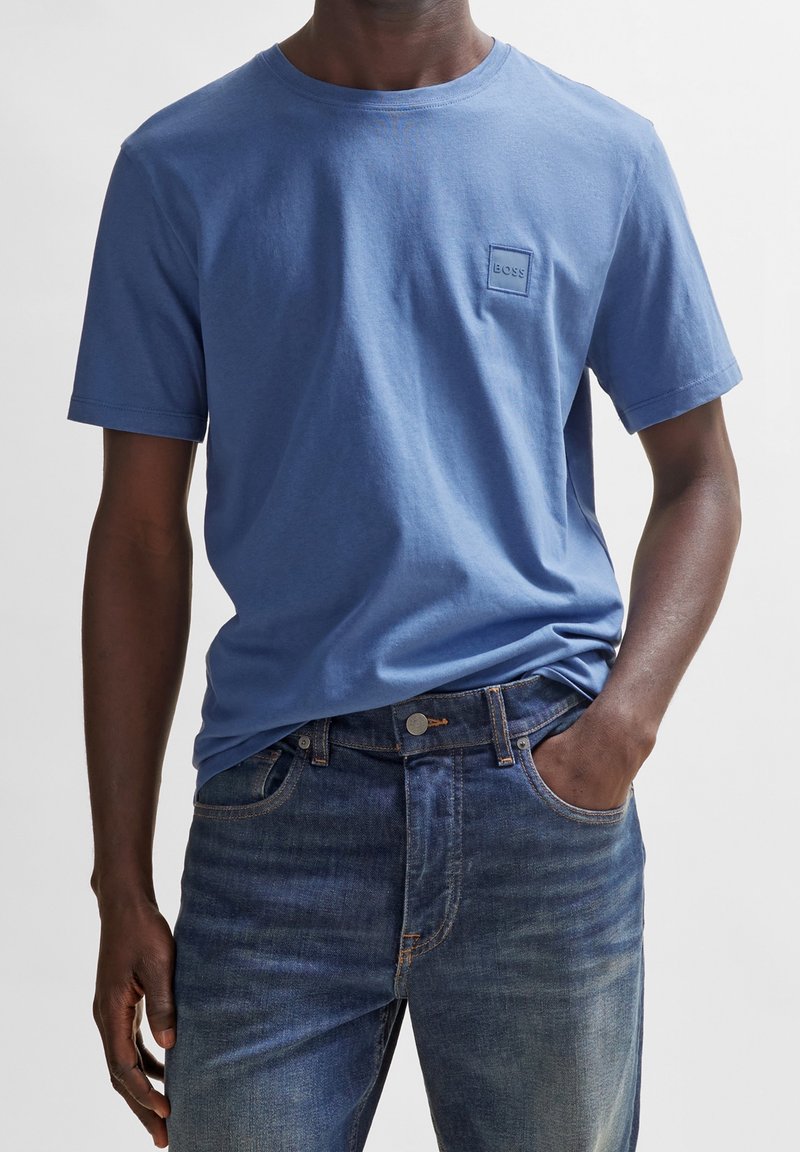 Homme portant un T-shirt bleu uni à manches courtes avec un petit logo "BOSS" sur la poitrine et un jean bleu foncé, une main dans la poche.