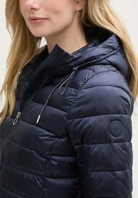Námornícka pufrová bunda s prešívaným dizajnom, má kapucňu, zipsové zapínanie, nastaviteľné šnúrky a kruhové logo na ľavom rukáve.