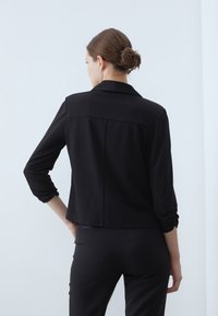 Schwarze, leichte Jacke mit langen Ärmeln, umgeschlagenen Manschetten und einer dezenten Rückennaht. Schlichtes, tailliertes Design ohne Verzierungen.