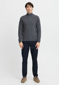 Grå ullgenser med høy krage og halv glidelås, kombinert med mørke jeans og lysebrune loafers. Modell som står mot en ensfarget bakgrunn.