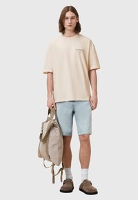 Béžové oversize tričko, světle modré džíny, šedohnědé obuv a nese světle hnědý batoh s popruhy a detaily v kontrastních barvách.