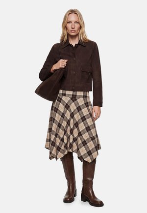 Kobieta ubrana w ciemnobrązową kurtkę, beżowo-brązową kratowaną spódnicę, wysokie brązowe buty i nosząca ciemnobrązową torbę na ramię.