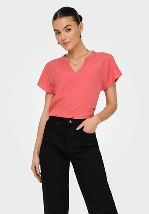 Woman with dark hair in a low bun wearing a coral short-sleeve top with a V-notch neckline and black high-waisted pants, hand in pocket.