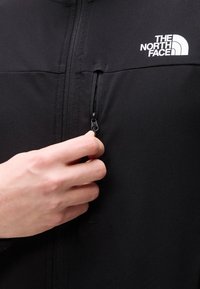 Χέρι που τραβάει το φερμουάρ σε μαύρο μπουφάν The North Face με λογότυπο στο στήθος, κοντινή λήψη στην υφή του υφάσματος και στη λεπτομέρεια του φερμουάρ.