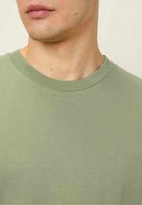 Gaiši zaļš kokvilnas T-krekls ar ribotu kakla izgriezumu, gludu tekstūru un pieguļošu formu, akcentējot augšējo ķermeņa daļu.