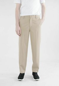 Beige Stoffhose mit geradem Schnitt, Gürtelschlaufen, einem Knopfverschluss und seitlichen Taschen; kombiniert mit einem schlichten weißen T-Shirt und schwarzen Turnschuhen.