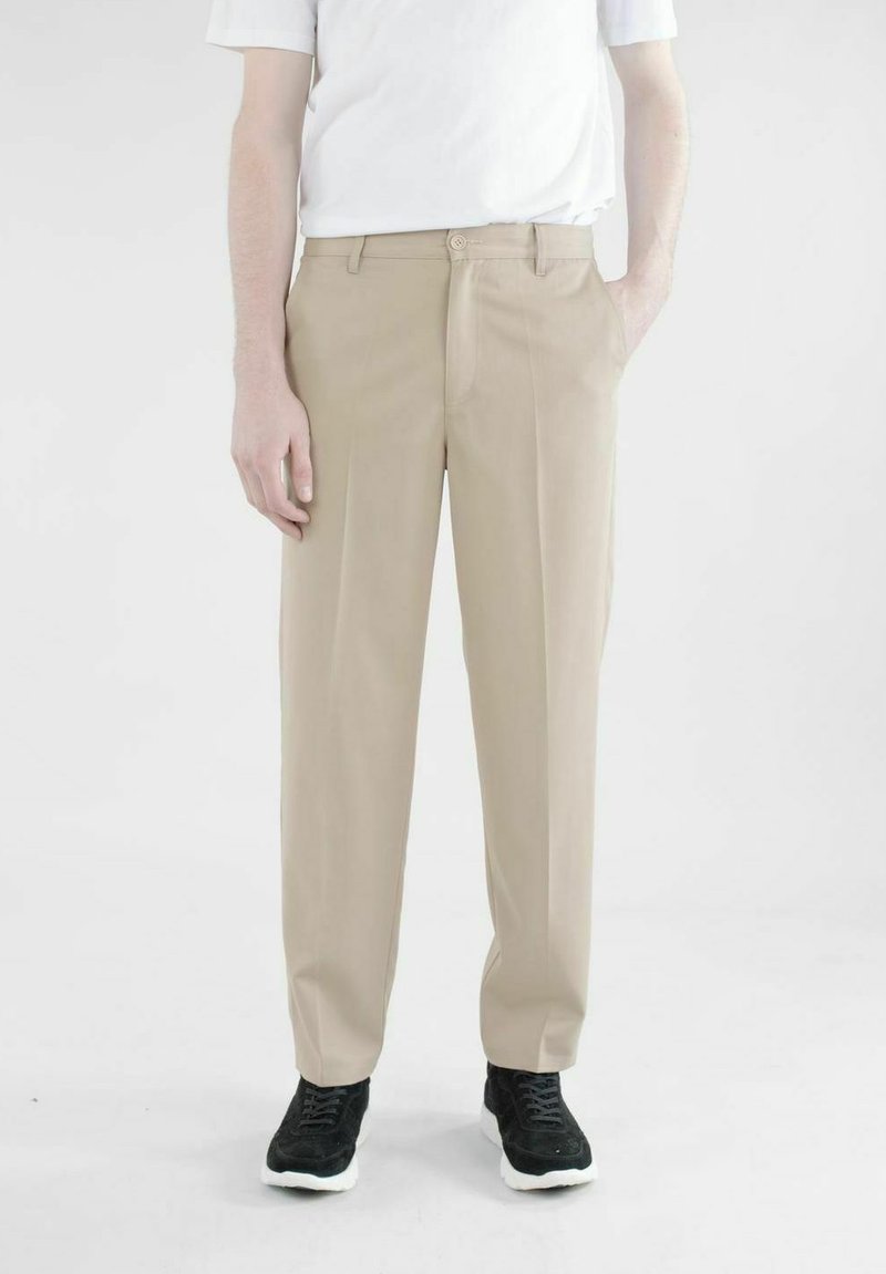 Beige Stoffhose mit geradem Schnitt, Gürtelschlaufen, einem Knopfverschluss und seitlichen Taschen; kombiniert mit einem schlichten weißen T-Shirt und schwarzen Turnschuhen.