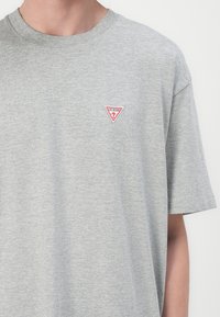 Graues Baumwoll-T-Shirt mit kurzen Ärmeln und rundem Ausschnitt. Verfügt über ein rotes und weißes dreieckiges Logo auf der linken Brustseite.